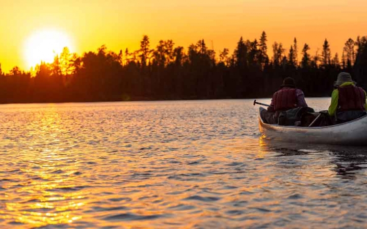 a canoe is paddled by two students while the sun sets behind a line of trees on the shore